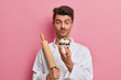 Male chef confectioner holds handmade delicious cake decorated with fresh berries, rolling pin, gives master class how to make yummy confectionery isolated on pink wall. Culinary, bakery, food concept