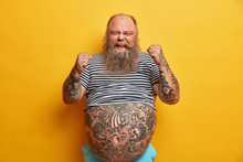 Joyful Bearded Man With Fat Belly, Clenches Fists, Happy As Won Competition, Has Tattoos, Dressed In Undersized Striped Shirt Feels Lucky Poses Indoor Over Yellow Background. Success, Winning Concept