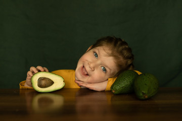 Close up photo of a beautiful girl with blue eyes and blonde hair, dressed in yellow, with half an avocado in her hand, next to her lies a whole avocado.Health and beauty concept.Dark green backgroun