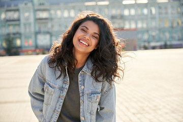 happy african young woman wearing denim jacket laughing looking at camera standing on street. smilin
