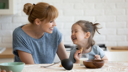 Funny lunch. Overjoyed laughing millennial mommy and her cute little daughter preschooler joking and having fun together while eating fresh homemade cookie with milk for snack at kitchen indoor