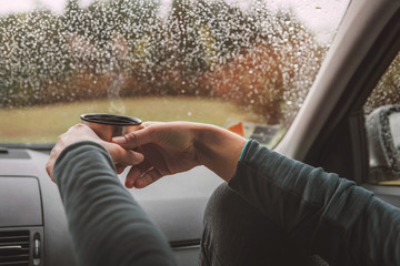 Wall Mural - Close up shot of graceful female palms holding the hot tea tourist thermos mug. She sitting on co-driver seat inside modern car, enjoying the moody rainy day weather looking through windscreen drops.