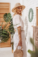 Young Elegant Fashionable Lady Wearing Summer White Crochet Jumpsuit, Straw Hat, Holding Wicker Bag, Posing At Home, In Stylish Boho Interior With Green Tropical Plants, Trendy Decorations 