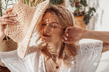 Happy smiling woman enjoying sunlight, hiding her face with straw hat, wearing trendy necklace with many chains, posing at home, in stylish boho interior. Summer fashion conception