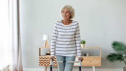 Wall Mural - Happy middle aged senior hoary retired woman using walking frame, enjoying rehabilitation procedures indoors. Smiling older disabled grandmother making steps with walker, handicapped people lifestyle.