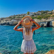© Руслан Полуэктов - A thin nice girl in a straw hat and a white dress stands on a rock and looks down at the bay with blue sea water. Beautiful woman on a background of Mediterranean cliffs relaxing by the sea. Hot day