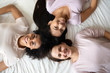© fizkes - Happy best girlfriend diverse ethnicity girls lying down in bed smiling looking at camera, three attractive women wearing comfy fashionable sleepwear awaken after sleepover posing in bedroom top view