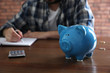 © New Africa - Man counting money at wooden table, focus on piggy bank