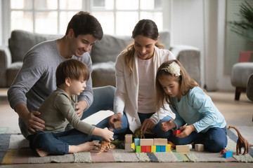 Canvas Print - Happy couple parents playing with small preschool children on warm floor in living room. Joyful family of four enjoying spending weekend free leisure time together, constructing building with cubes.