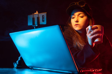 Poster - Freelancer girl with a glass. Woman in a dark room with a computer. Freelancer with a laptop on a black background. Pensive freelancer girl in cap. Woman performs doing remote job via laptop computer