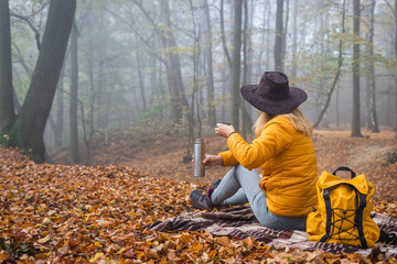 Wall Mural - hiker sitting on picnic blanket and drinking hot drink in autumn forest. woman resting in nature dur