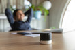 © fizkes - Close up focus on portable stylish little digital assistant on table, with relaxed businesswoman on background. Happy worker using small voice speaker or enjoying pleasant music from column in office.