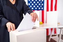 USA. US Elections. Woman Tosses Election Bill Into A Box. Human Voter On Background Of USA National Flag. American Woman At Polling Station. American In Presidential Election. Chooses President