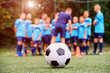 © Serhii - Soccer ball and blurred youth soccer team with coach on the field, young players listening to coaching strategy of the game before training match. Selective focus