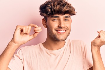 Young hispanic man holding macaron screaming proud, celebrating victory and success very excited with raised arm