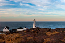 Lighthouse Cylinder Shape Concrete Structure On A Red Rocky Cliff With Multiple Small White Outcrop Buildings. The Cloudy Red Sky Is Setting In The Background With The Blue Horizon And Ocean.