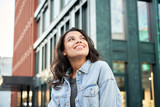 Fototapeta  - Confident happy beautiful young hipster African American woman wearing denim jacket looking up standing on city street outdoors dreaming, thinking or good future on urban buildings background.
