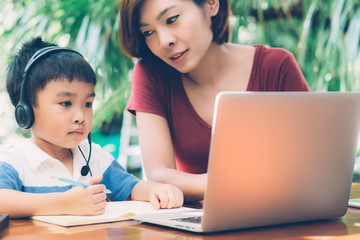 Wall Mural - Young asian mother and son using laptop computer for study and learning together at home, boy writing on notebook for homework and wearing headphone, teacher or mom support child, education concept.