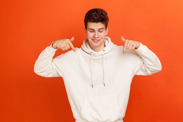 Smiling young man in casual style white hoodie pointing fingers down, showing on advertising area, free space for commercial. Indoor studio shot isolated on orange background