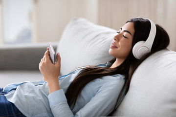 Wall Mural - Audio Books App. Asian Woman With Smartphone And Headphones Relaxing At Home