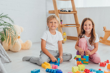 excited brother and sister in pajamas sitting on floor near building blocks and looking at camera