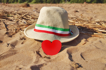 wooden scarlet heart and straw hat with ribbon with colors of flag of Italy green, white and red in golden sand on beach, selective focus