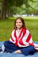 Wall Mural - Vertical full length portrait of cute girl covered by American flag sitting on picnic blanket in park and smiling at camera, copy space