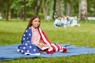 Wall Mural - Full length portrait of cute girl covered by American flag sitting on picnic blanket in park and smiling at camera, copy space