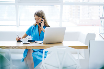 Busy stylish manager consulting clients via smartphone while noting some information in notebook.Beautiful female having mobile conversation while writing down text in notepad sitting at laptop device