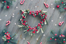 Decorative Heart Shaped Mistletoe Wreath. Wooden Christmas Background Decorated With Natural Fir Twigs, Red White Xmas Stripy Candy Canes And Red Berries.