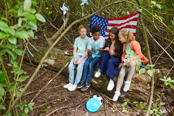 Wall Mural - Multi-ethnic group of kids using digital tablet while playing in yard hiding under bushes with American flag in background, copy space
