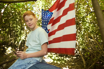 Portrait of red haired teenage boy looking at camera outdoors while hiding under branches of big tree in forest or playing in back yard with American flag in background