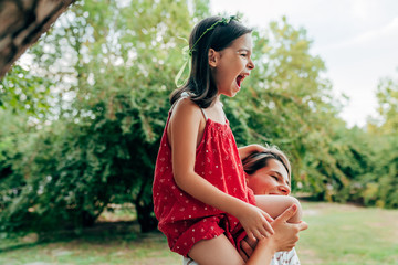 Wall Mural - Smiling mother playing and riding on her shoulder her daughter in the park. Happy kid having fun with her mom enjoying the time together. Mother and child share love with each other.