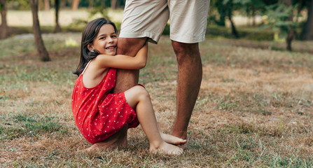 Wall Mural - Cheerful smiling daughter spending time with his father swinging on daddy's leg outdoors. Ñute little girl playing with her dad in the park. Daddy and kid having fun together Happy father's day.
