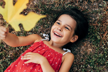 Wall Mural - Portrait of smiling little girl holding and looking in her hand a big yellow leaf hiding her face in the park. Kid exploring a big leaf during lying on the grass outdoors.