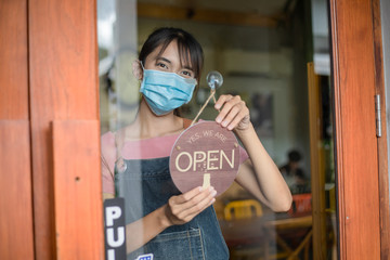 Asian woman barista owner turning sign to opening the coffee shop while wearing protective face mask