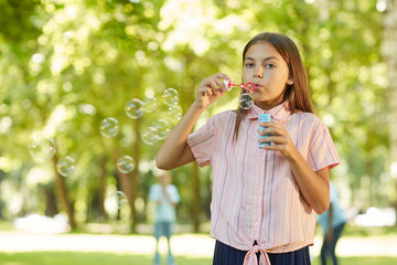 Wall Mural - Waist up portrait of teenage girl blowing bubbles and looking at camera while standing in green park outdoors, copy space
