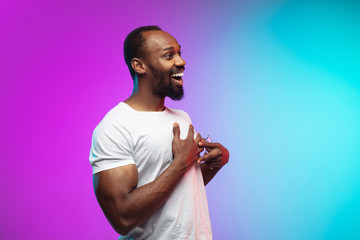 Wall Mural - Shy, happy, smiling. African-american young man's portrait on gradient studio background in neon. Beautiful male model in casual style, white shirt. Concept of human emotions, facial expression, sales