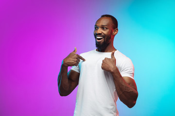 Wall Mural - Pointing, showing. African-american young man's portrait on gradient studio background in neon. Beautiful male model in casual style, white shirt. Concept of human emotions, facial expression, sales