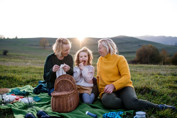 Small girl with mother and grandmother having picnic in nature at sunset.