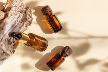 Glass cosmetic bottles with a dropper stand next to a log on a beige background with bright sunlight. The concept of natural cosmetics, natural essential oil