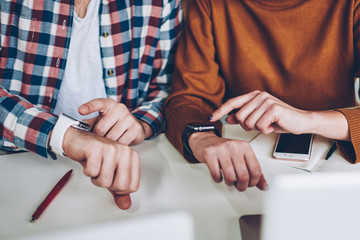 Cropped photo of male and female skilled freelancers checking notification at modern smartwatch in break of working with laptop computers, two students connected to gps tracking sitting indoors