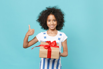 Smiling little african american kid girl 12-13 years old isolated on blue background. International Women's Day birthday, holiday concept. Hold red present box with gift ribbon bow, showing thumb up.