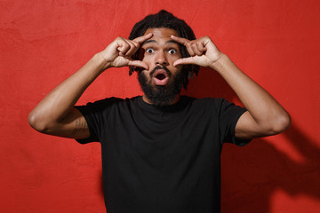 Shocked amazed young african american man guy with dreadlocks 20s in black casual t-shirt posing stretching eyelids, keeping mouth open looking camera isolated on red color background studio portrait.