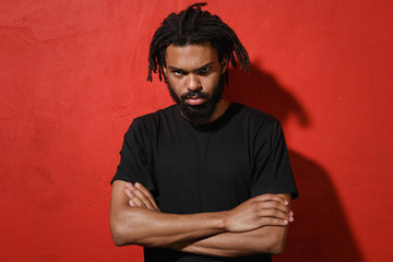 Wall Mural - Displeased offended concerned young african american man guy with dreadlocks 20s wearing casual black t-shirt posing holding hands crossed isolated on bright red color wall background studio portrait.
