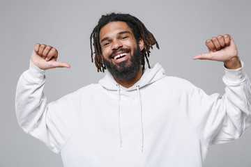 Cheerful young african american man guy with dreadlocks 20s wearing white casual streetwear hoodie posing pointing thumbs on himself looking camera isolated on grey color background studio portrait.