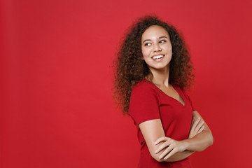 Wall Mural - Side view of smiling young african american woman girl in casual t-shirt posing isolated on red background studio. People lifestyle concept. Mock up copy space. Holding hands crossed, looking aside.