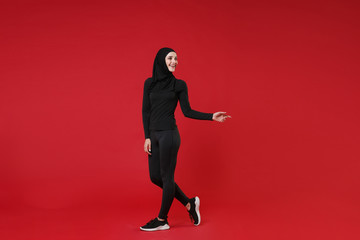 Full length portrait of smiling young arabian muslim woman in hijab black clothes isolated on red wall background. People religious lifestyle concept. Mock up copy space. Pointing index finger aside.