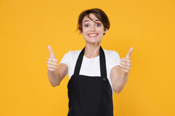 Smiling young female woman 20s barista bartender barman employee in white t-shirt apron work in coffee shop posing showing thumbs up looking camera isolated on yellow color background studio portrait.