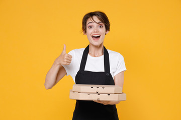 Excited young female woman 20s barista bartender barman employee in t-shirt apron holding italian pizza in cardboard flatbox showing thumb up isolated on yellow color wall background studio portrait.